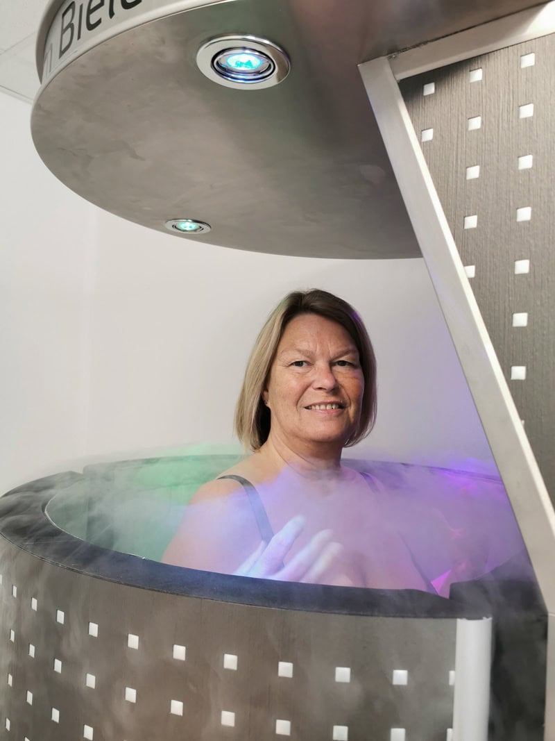 Eine lächelnde Frau in einer grauen Kryosauna mit Nebel. Hintergrund weiß und Beleuchtung farbig.