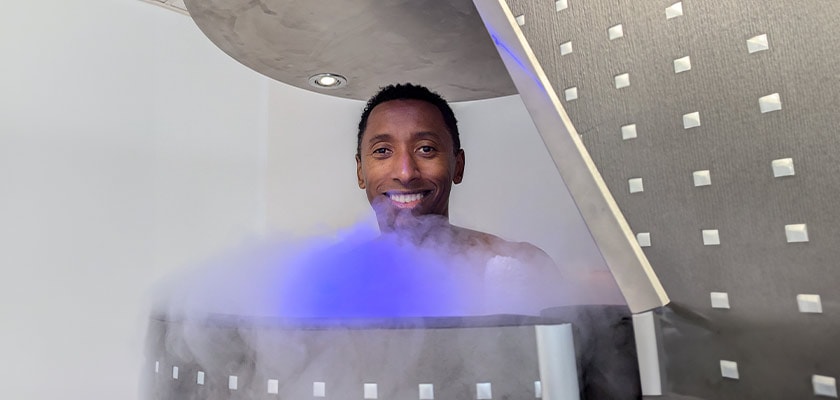 Ein lächelnder Mann in einer Kryokammer mit blauem Nebel im Vordergrund.