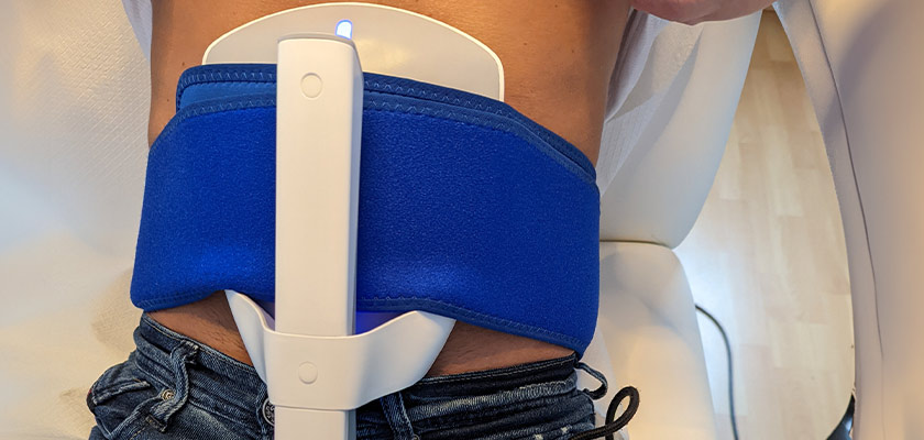 Eine Person trägt ein blaues medizinisches Gerät am Bauch auf hellbraunem Sofa.