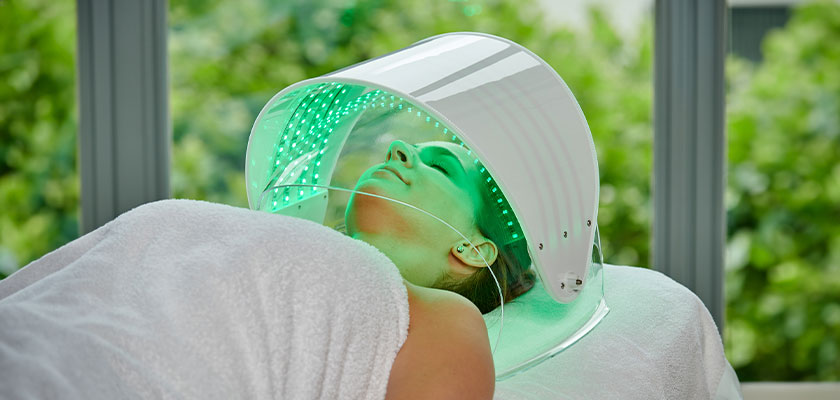 Person unter LED-Maske mit grüner Beleuchtung, entspannt. Weiße Decke, Hintergrund grün.