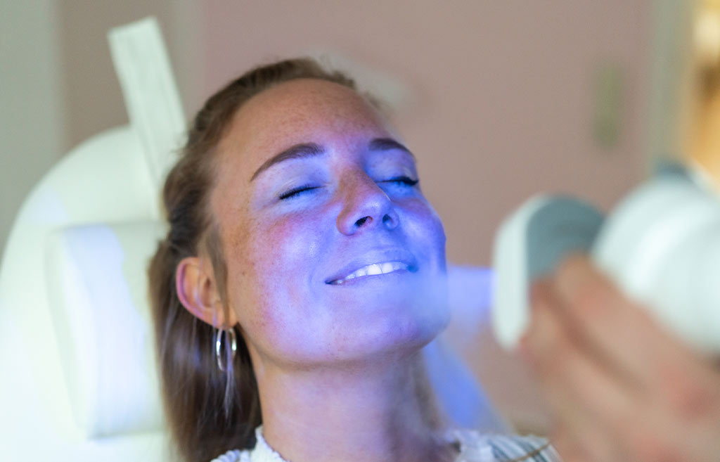 Eine lächelnde Frau unterzieht sich einer Gesichtslichtbehandlung, blauer Lichtschimmer.